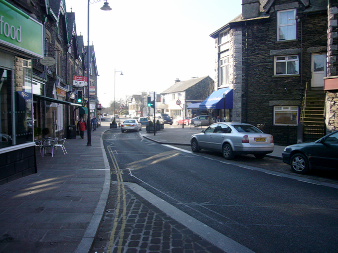 Cars parked on town street with wide pavement next to a cafe. 