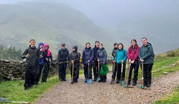 Group of young people in waterproofs standing together on track. They are holding spades and stand in front of mountains obscured by the rain