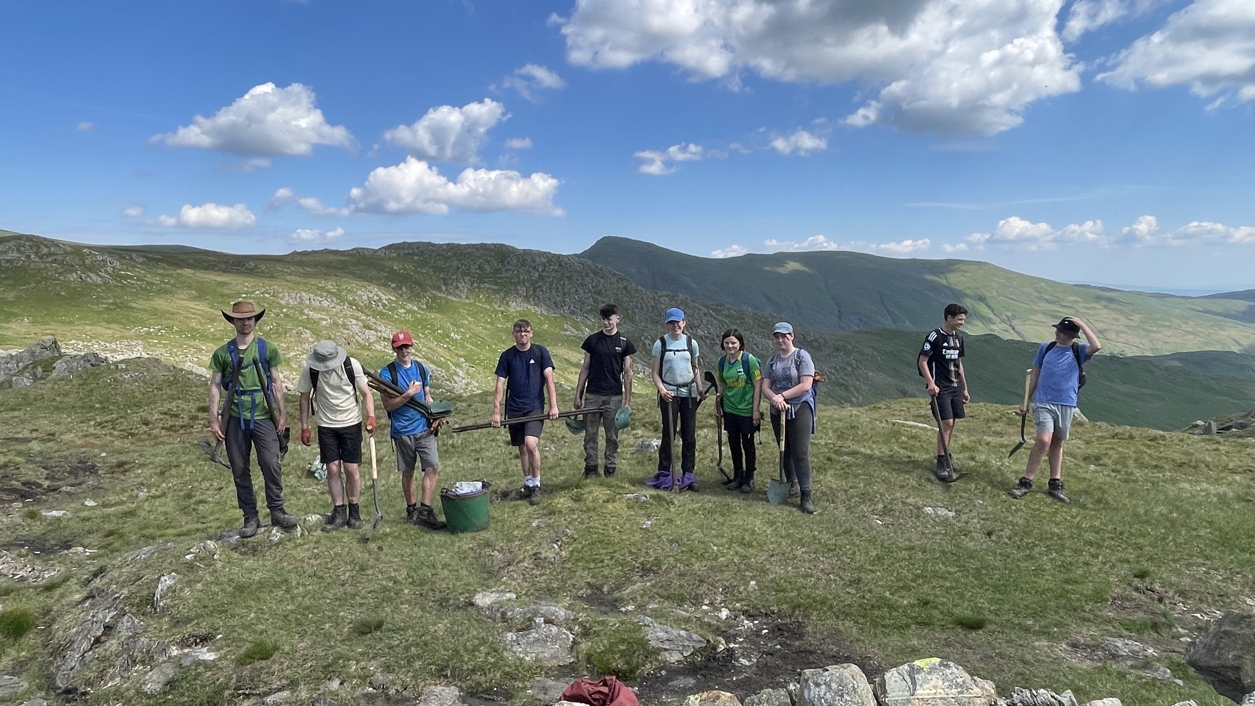 Group of young people in outdoor gear, holding buckets and spades. They are standing on a grassy fell in sunshine with mountains in background.