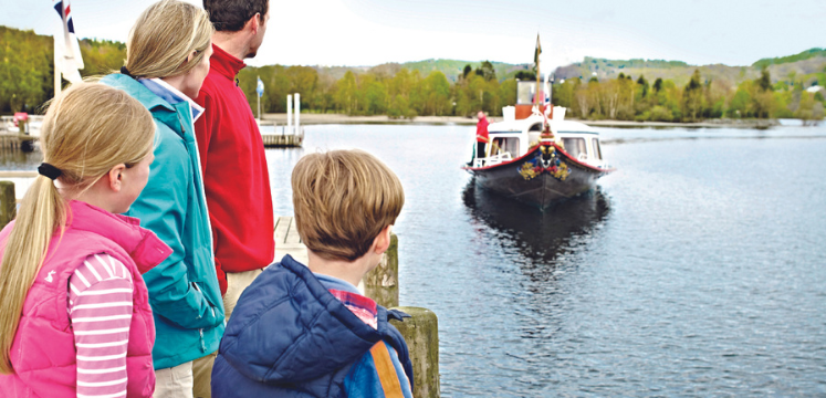 A family look on to COnsiton Water with a boat sailing towards them 