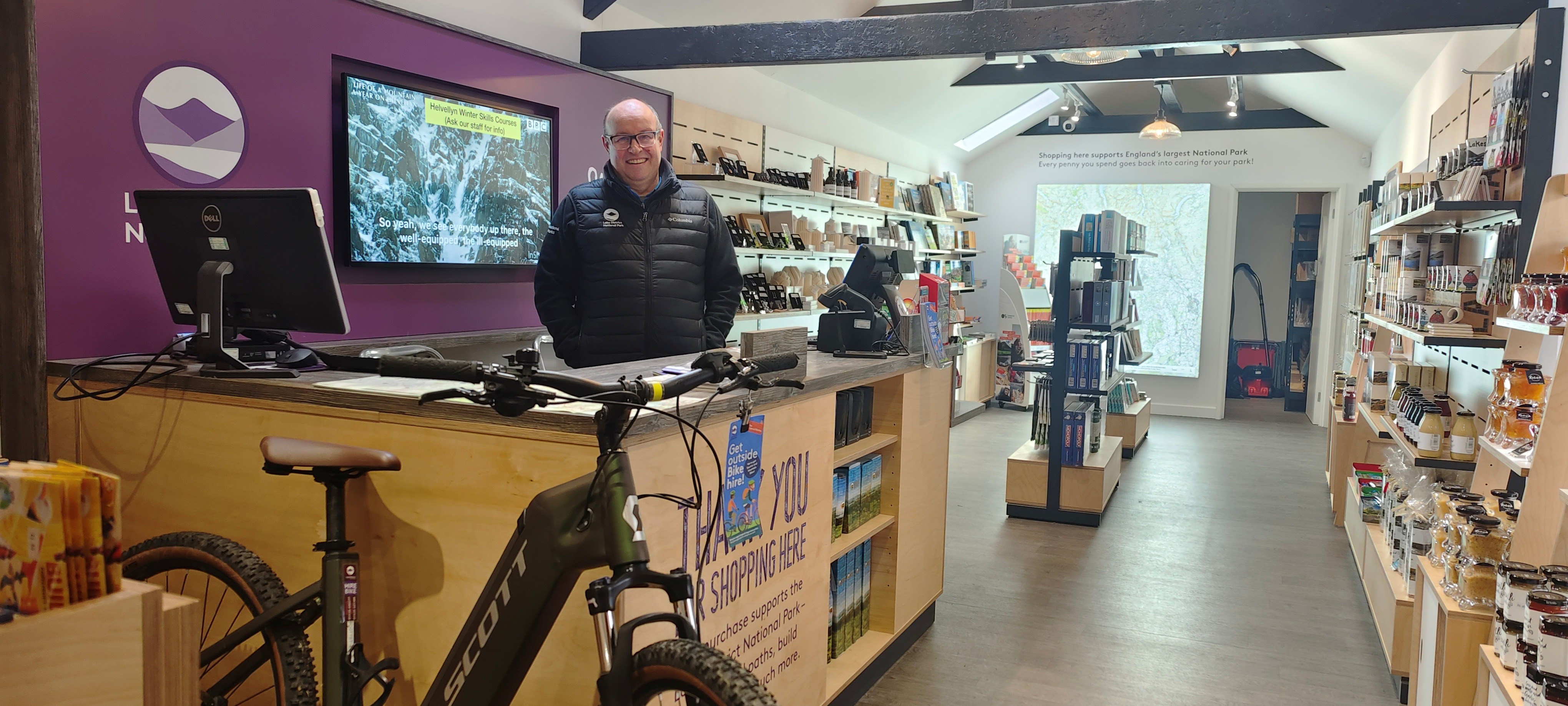 Andy Pearce stands behind the till at Hawkshead Information Centre in front of a purple background with the Lake District National Park logo and World Heritage Site logo