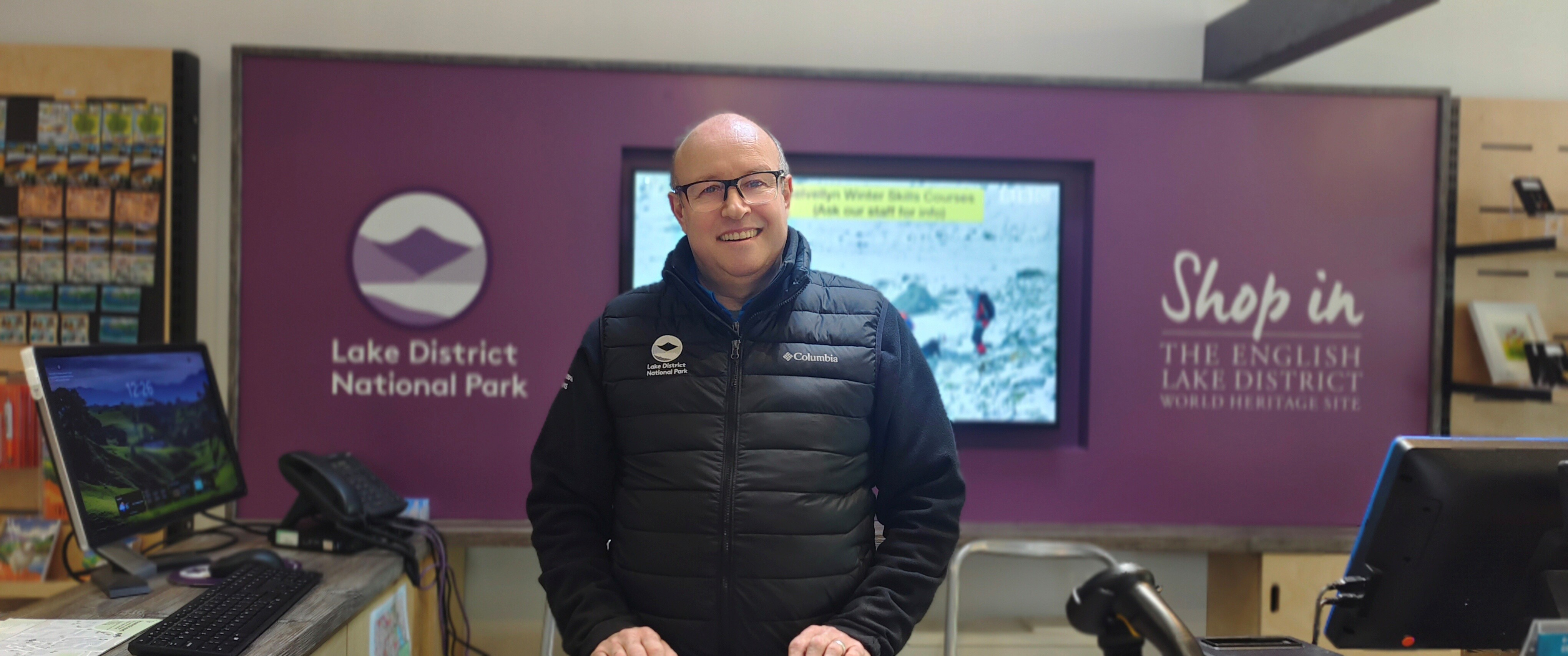 A man stands behind the counter at Hawkshead Information Centre in front of a purple background which has the Lake District National Park logo