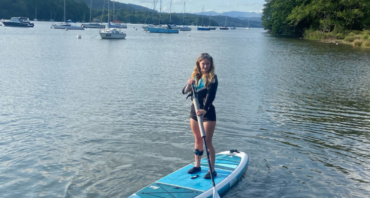 Young woman, Amelia, stood on white and blue paddleboard on the lake. She is wearing black shorts, a top and a lifejacket.