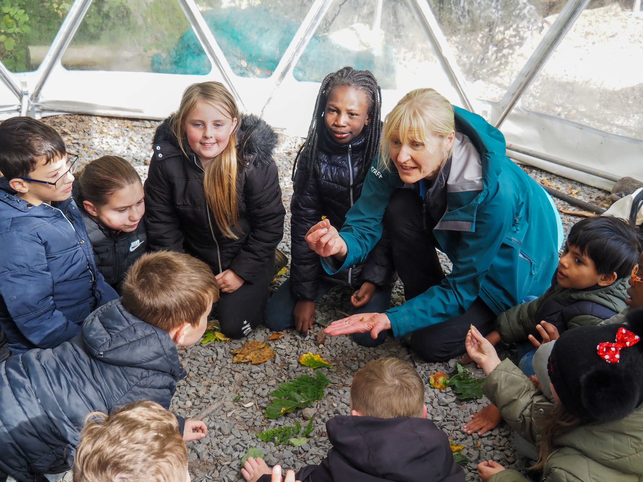 A group of children crouching on the ground with Learning and Engagement Manager Ruth Suddaby, as they listen to her intently