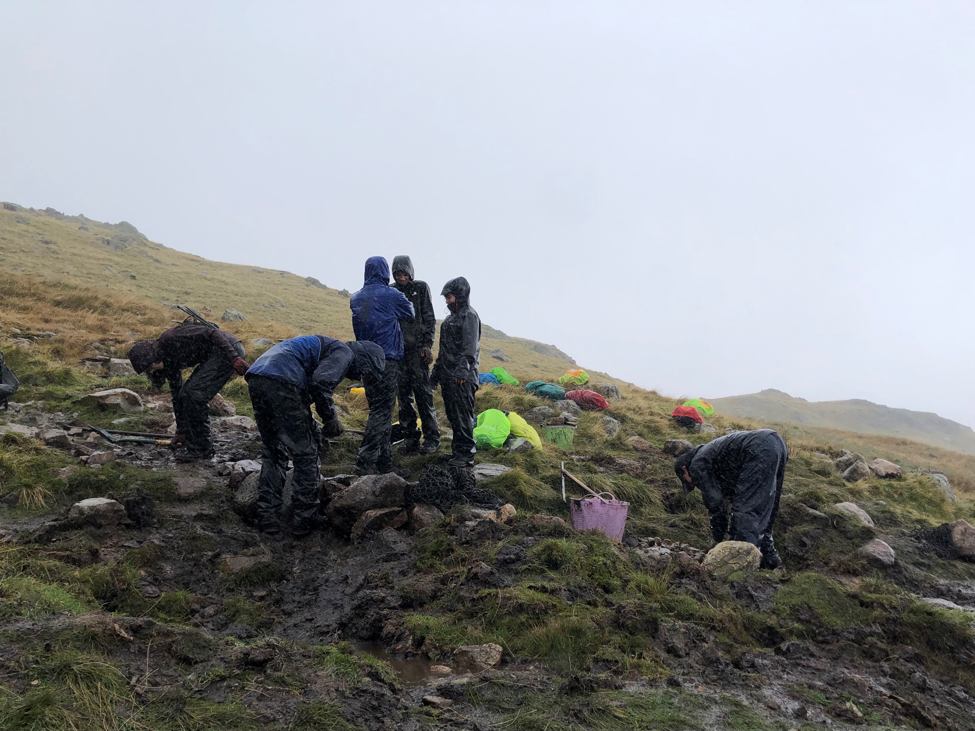 Group of young people in waterproofs working with spades and tools to fix footpaths. They are spread out over a rocky footpath on a grassy fell and distant fells are obscured by rain.