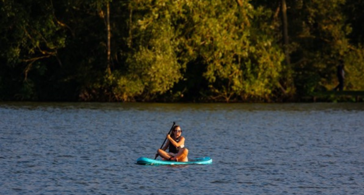 Woman sat cross-legged on a blue paddleboard on a lake with green trees behind