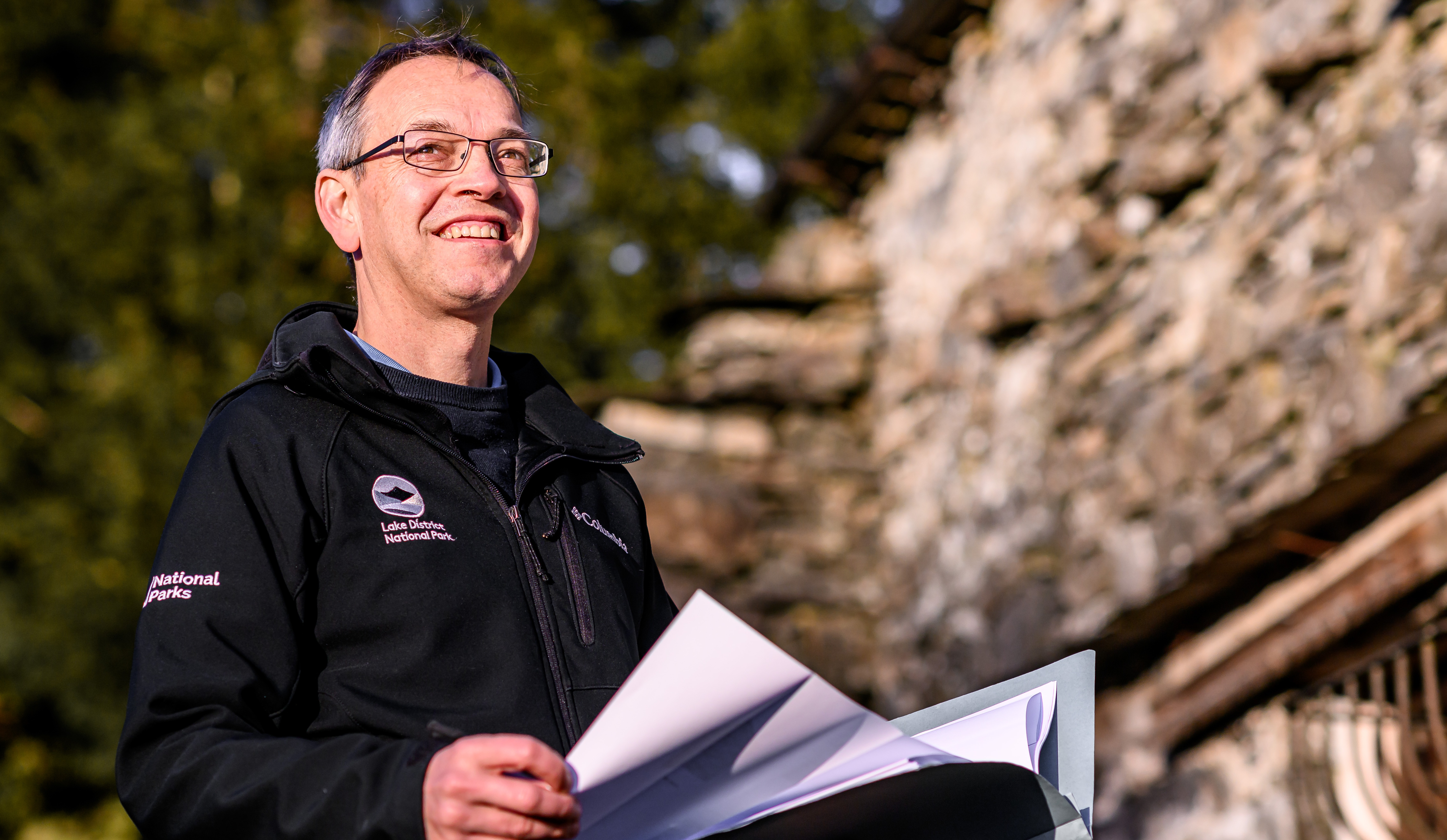 Male Planner standing outside a traditional lakeland building. He is wearing a black uniform jacket which show the LDNPA and National parks logos.