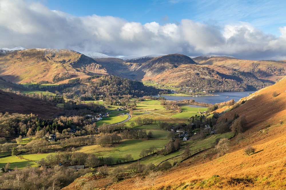 aerial view of ullswater and patterdale on a clear sunny day, with rolling fells in hues of orange, green and brown below a cloudy blue sky