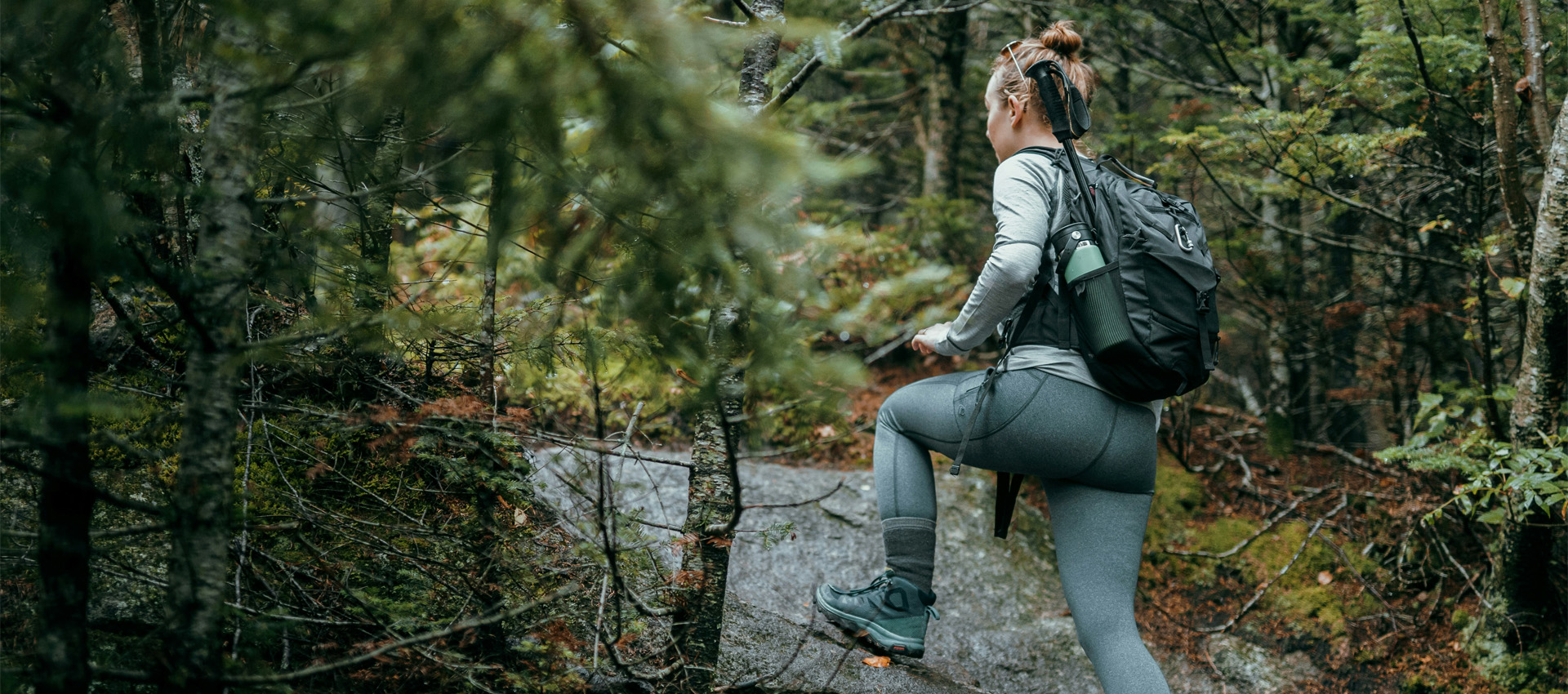 white adult female in grey walking clothing stepping on a stone path amongst dark green woodland