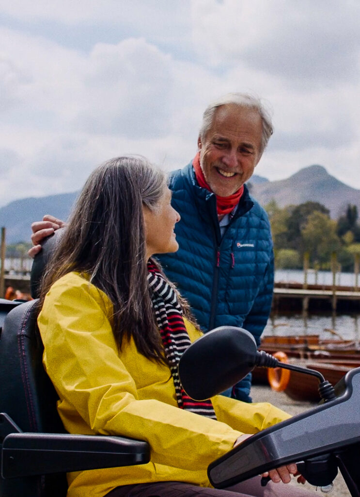 A couple, including a woman in a yellow jacket using a mobility scooter beside a lake