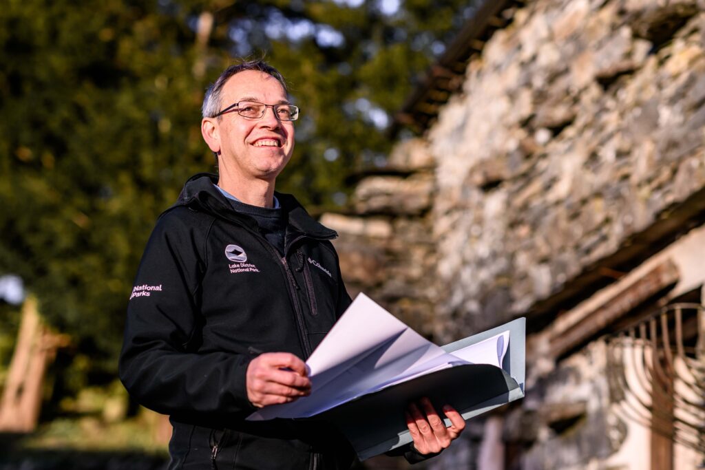 white adult male in a black Lake District National Park jacket holding open documents outdoors near a stone building