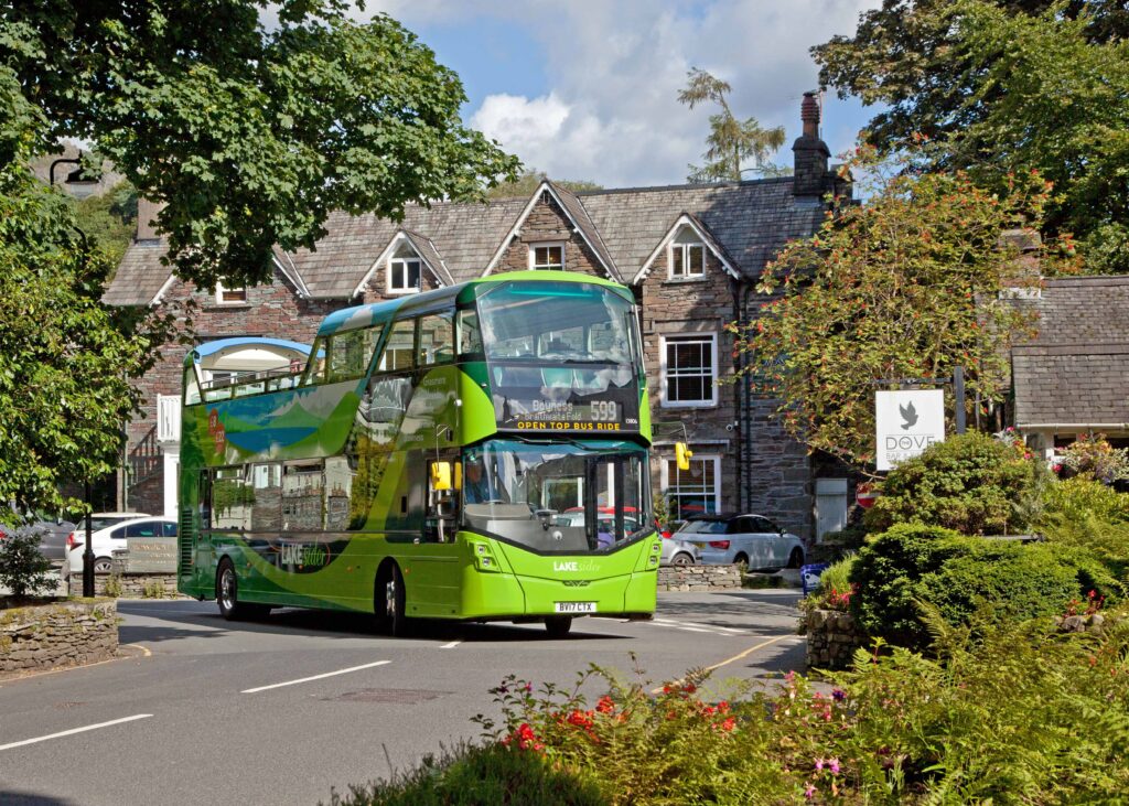 Green double decker bus in Grasmere on road in front of stone country building. Sunny day