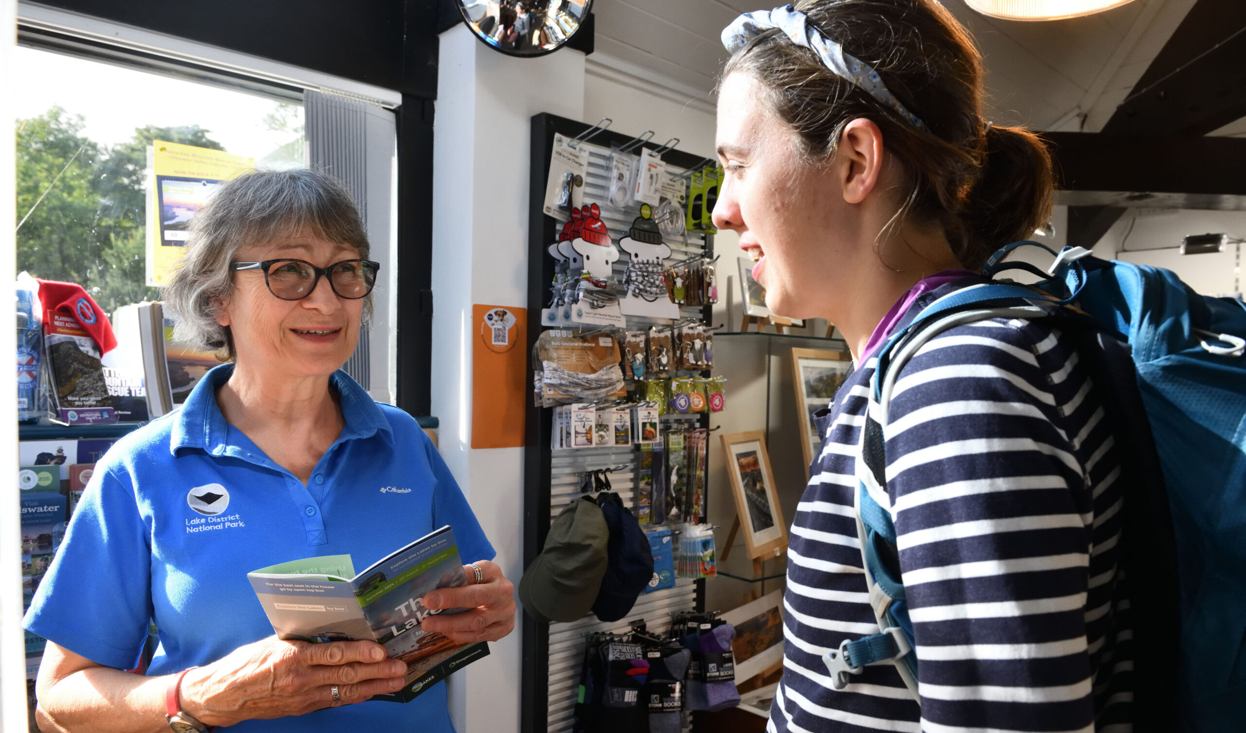 Customer Adviser staff member holding leaflets and speaking with a person wearing a backpack inside a Lake District National Park information centre.
