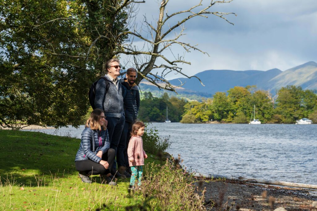 child, adult female, adult male, elderly male, stood together on grass lake front under green tree, looking out over a lake on a sunny day