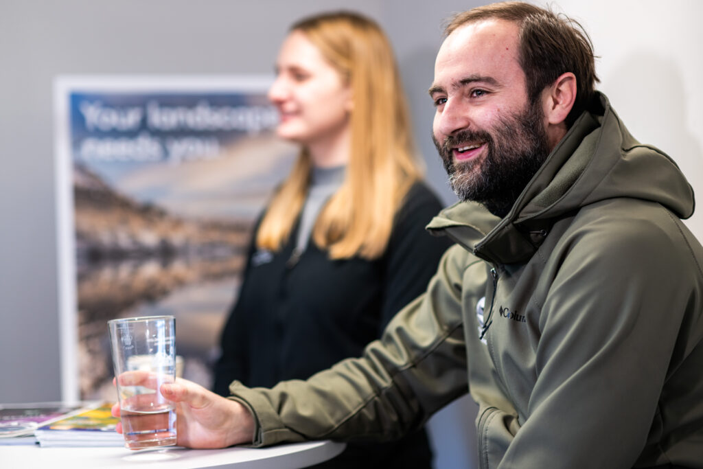 adult male and adult female in lake district national park authority uniform sat smiling in office room