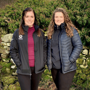 Two women standing outdoors in front of a stone wall and greenery, wearing outdoor jackets.