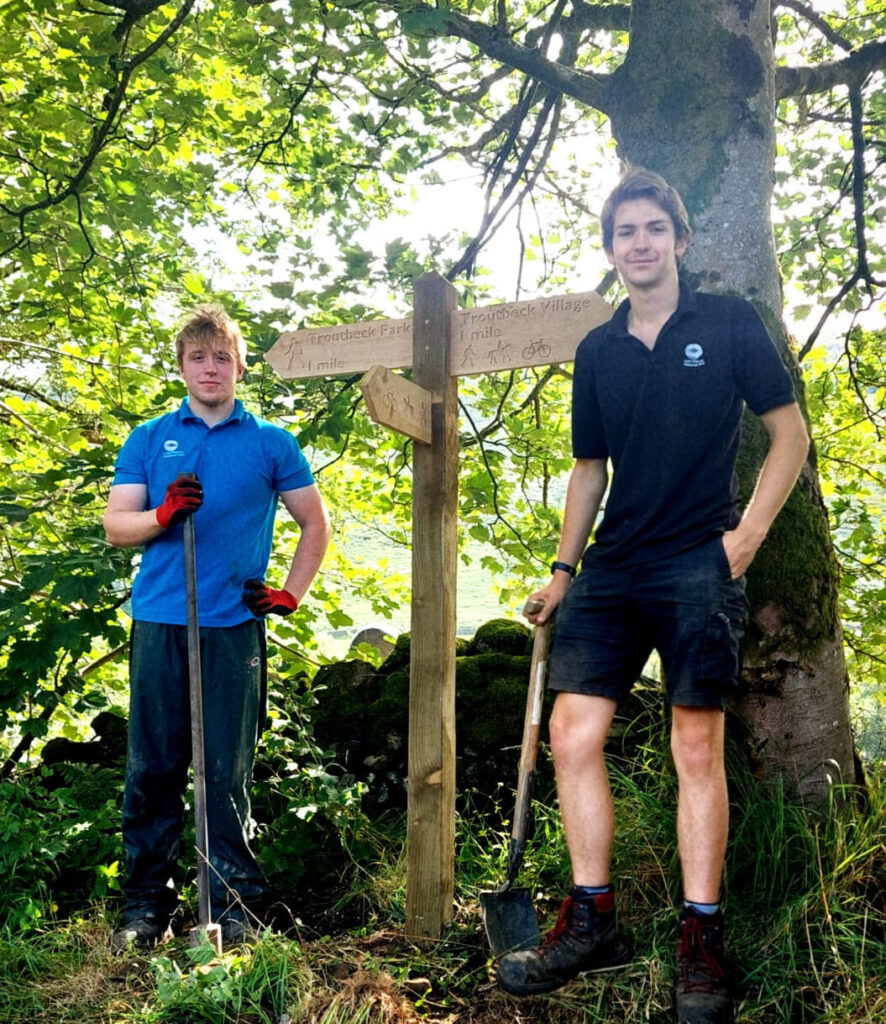 Two males outdoors installing a wooden signpost near a tree, holding spades
