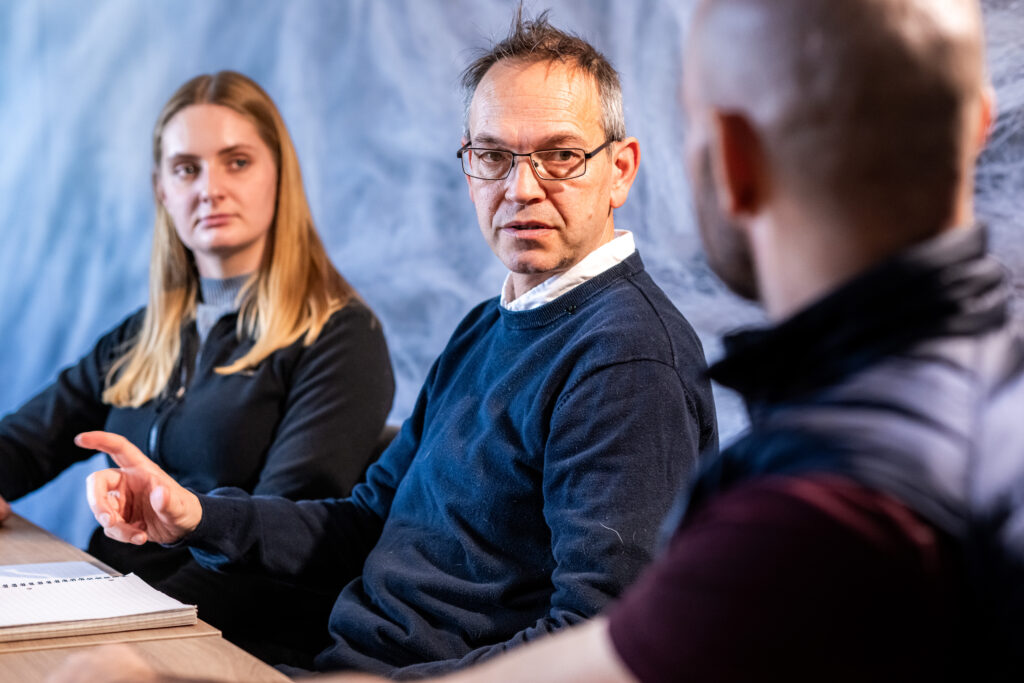 Three people sitting at a table in a meeting room, engaged in discussion with a notebook on the table.