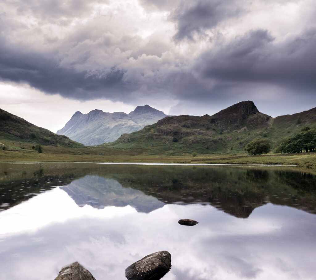 An image of the water on Blea Tarn, with fells reflected in the water on the horizon, and looming grey clouds