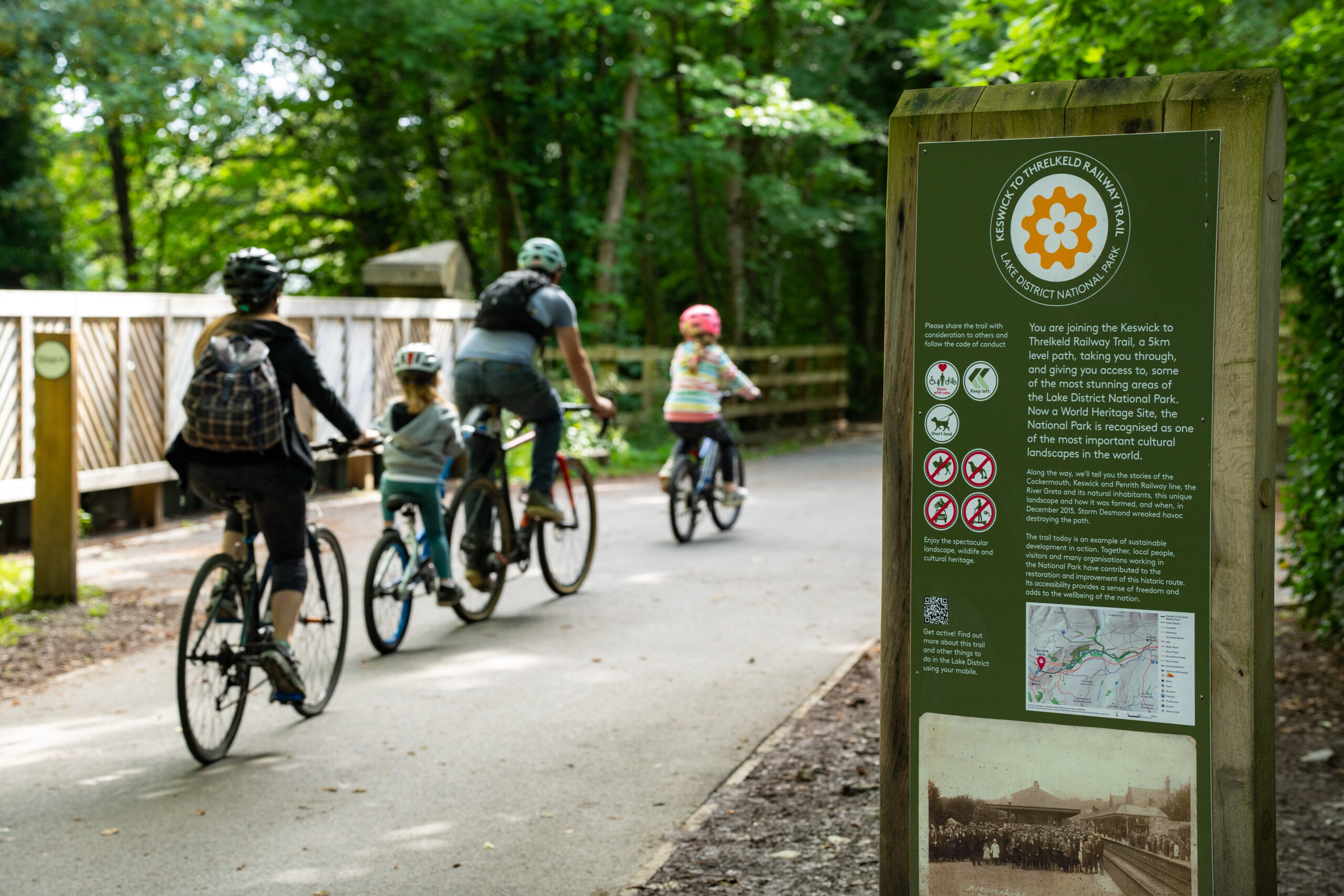 A family, mum, dad and two young girls, riding cycles along the Keswick Railway path with a greeninterpretation panel at the side of the path