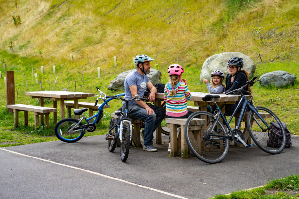 Family wearing helmets with a young girl sat on picnic benches with the bikes resting up against them