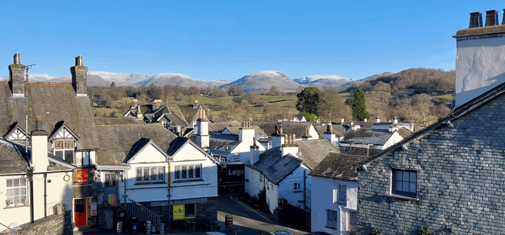 high up view of villlage with white houses with slate roofs on a sunny day with snow capped fells in backdrop
