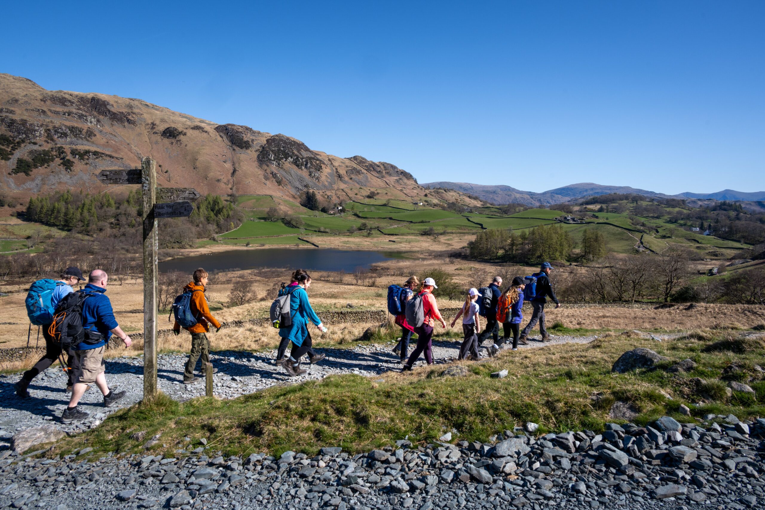 Group of people walking on a path with blue skies and a tarn in the background, on a Lake District National Park Guided Walk.