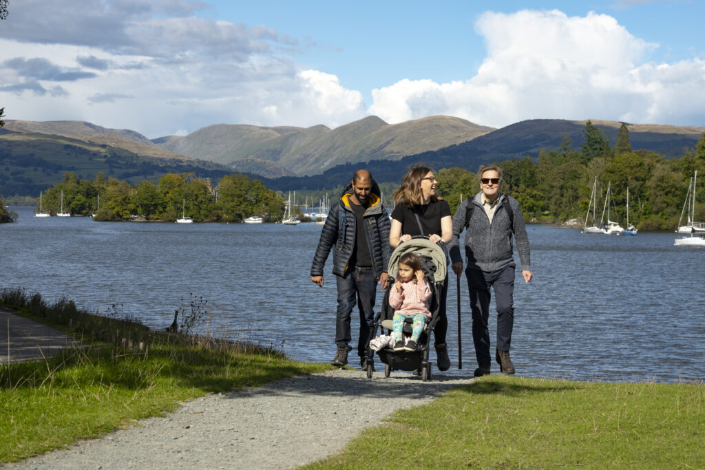 Young family, mum pushing pushchair and dad and grandad walking alongside on a gravelled path with Windermere lake and mountains behind