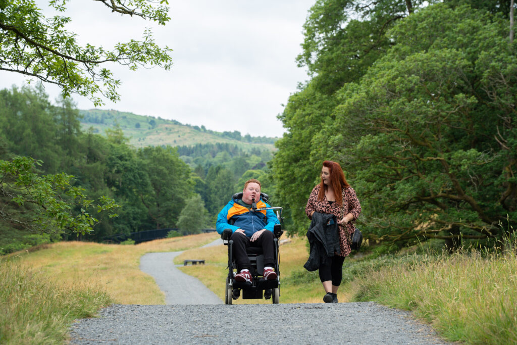 A women walking a alongside a man in a electric wheelchair on a gravelled path surrounded by large trees