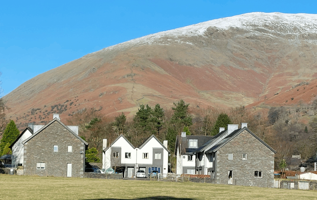 group of white and stone rural village buildings in front of a tall barren red and brown snow dusted fell on a sunny day