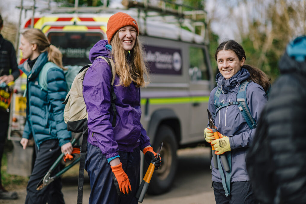 Two teenage girls in outdoor clothing smiling in front of a Lake District Landrover on a Young Rangers volunteer day