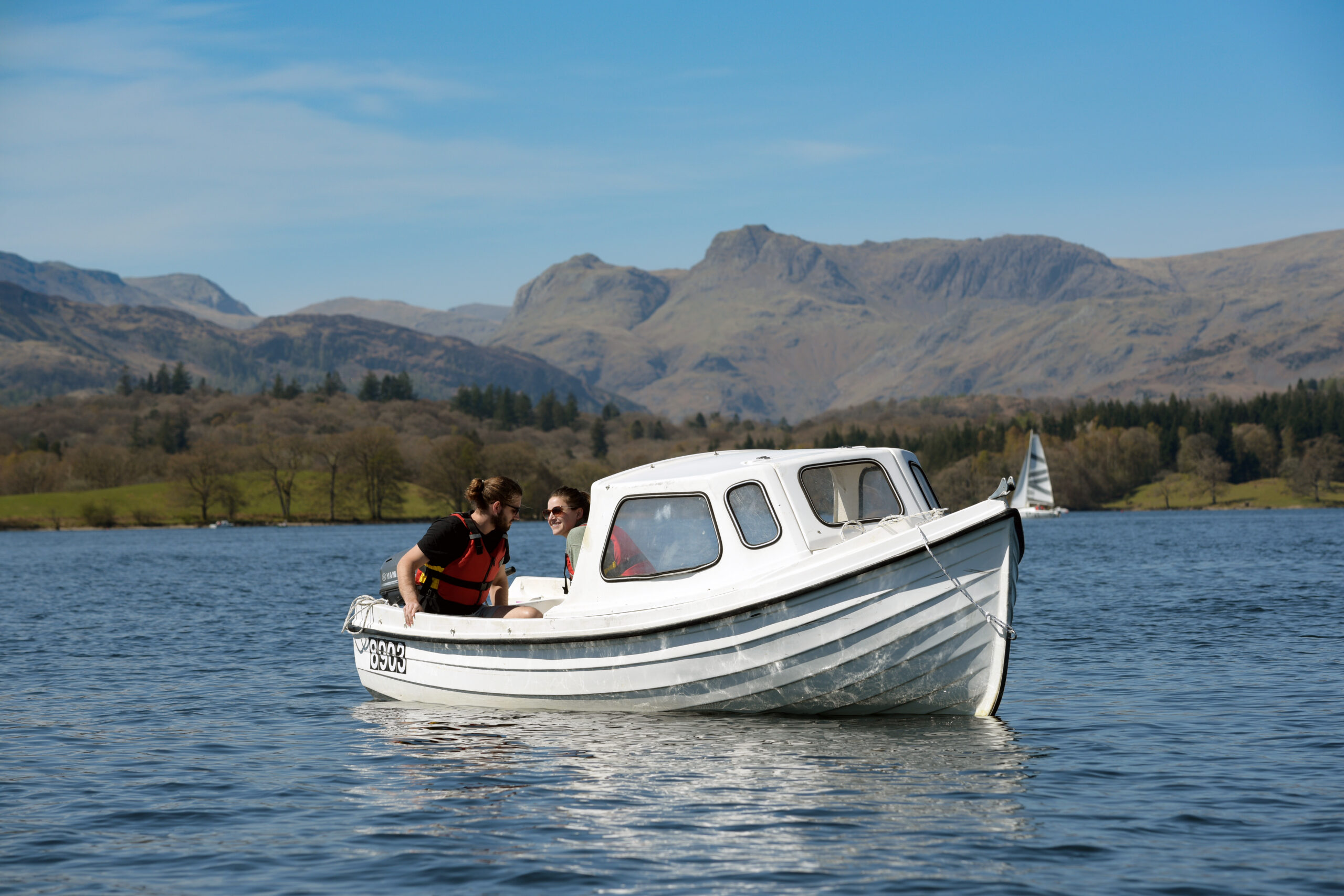 Small white motorised boat on lake, with fells in the background on a blue sky sunny day