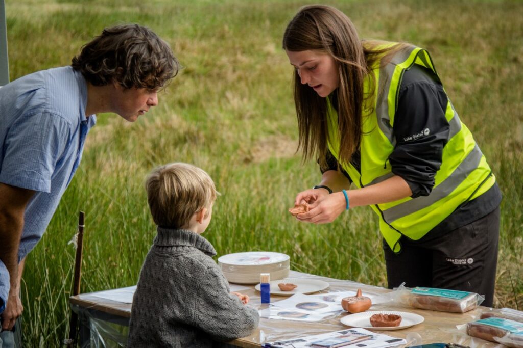 adult male and child at table outdoors with an adult female in high vis showing them an archaeological artefact in her hands