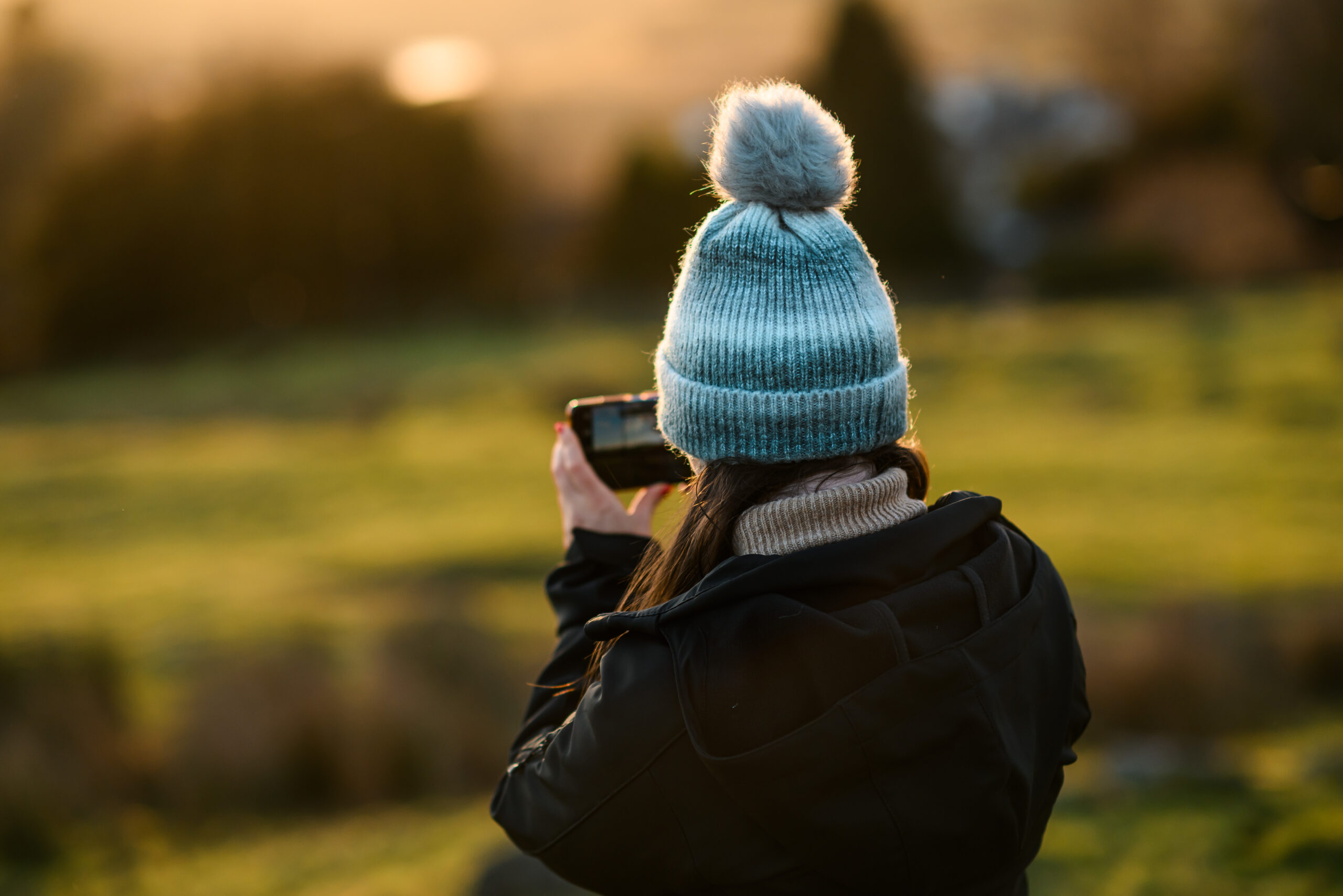 Adult woman from behind, wearing a black coat and light blue knit bobble hat, holding a photo taking a photo. Background is blurred, showing glowing green fields and brown trees