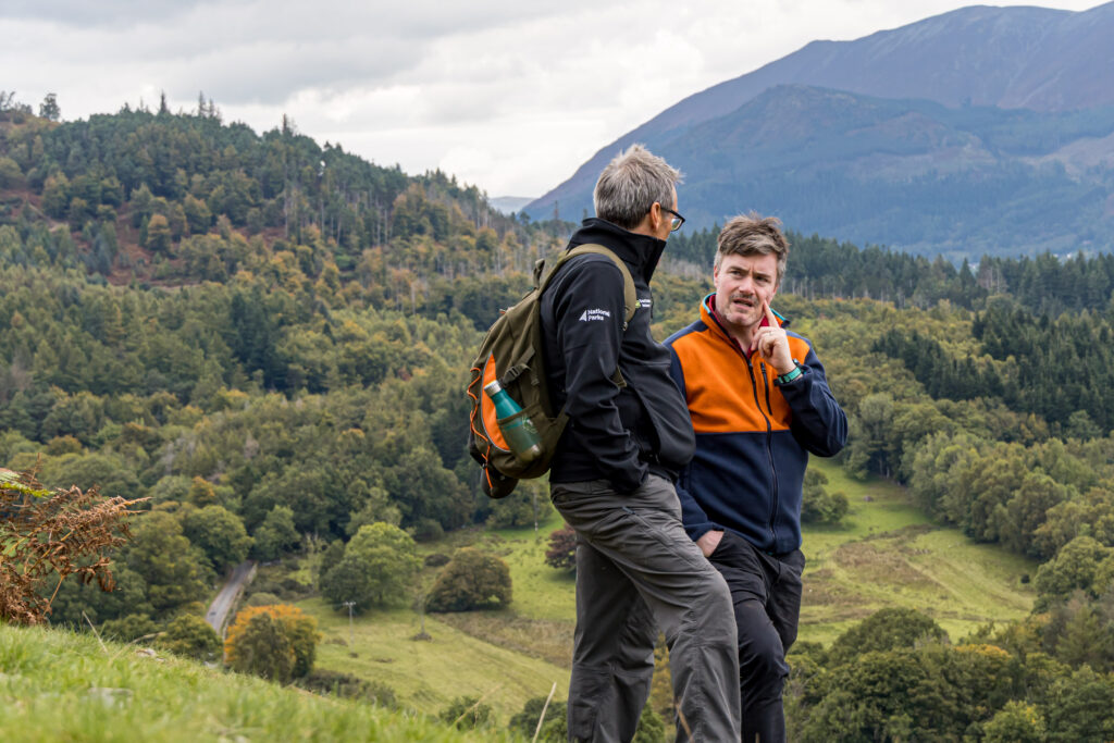 two white adult males stood talking in in front of fell and woodland backdrop