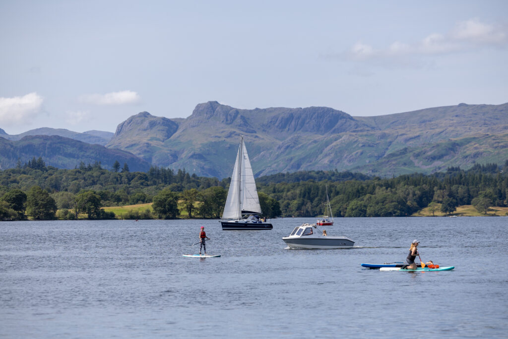 Windermere with a paddleboarder, kayaker and a small white motorised boat and sailing boat on the calm water. wooded lakeshore and fells in in the distance. blue sky with some clouds