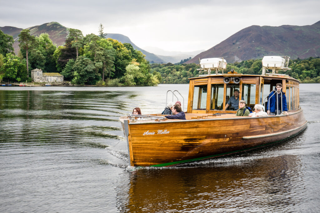 passengers on wooden boat cruise on Derwentwater. Grey sky and lake, green woodland and fells in backdrop