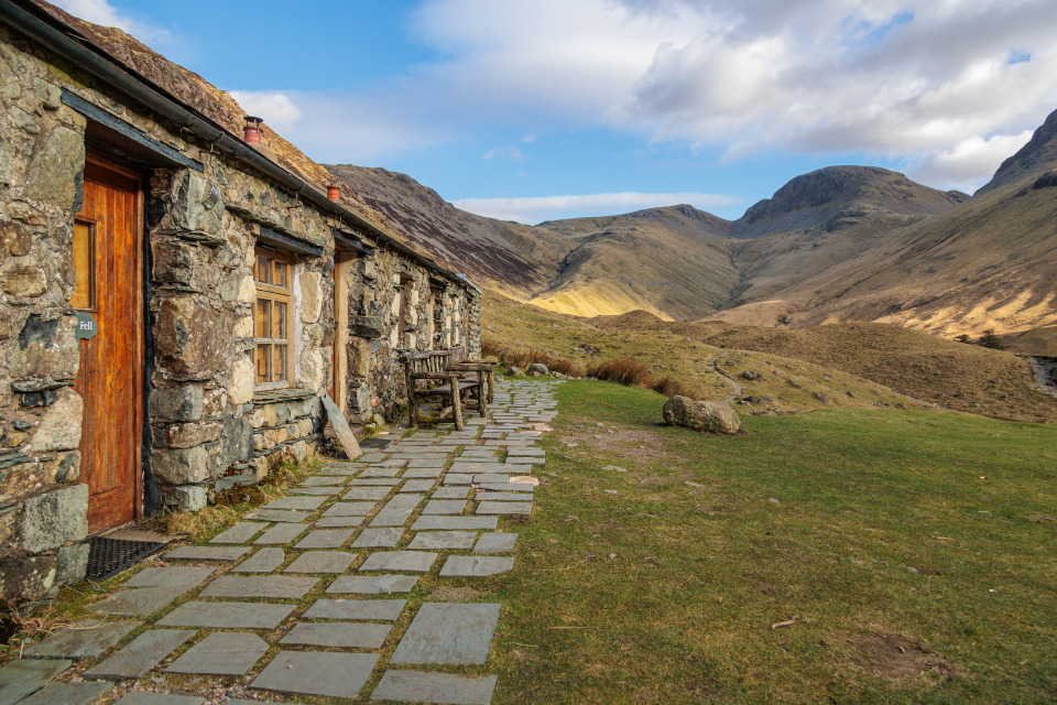 Black Sail Youth Hostel. Single storey old stone building with wooden doors and windows, lined by a stone path in front. Hostel overlooks views green, brown and grey fells. Blue partly cloudy sky