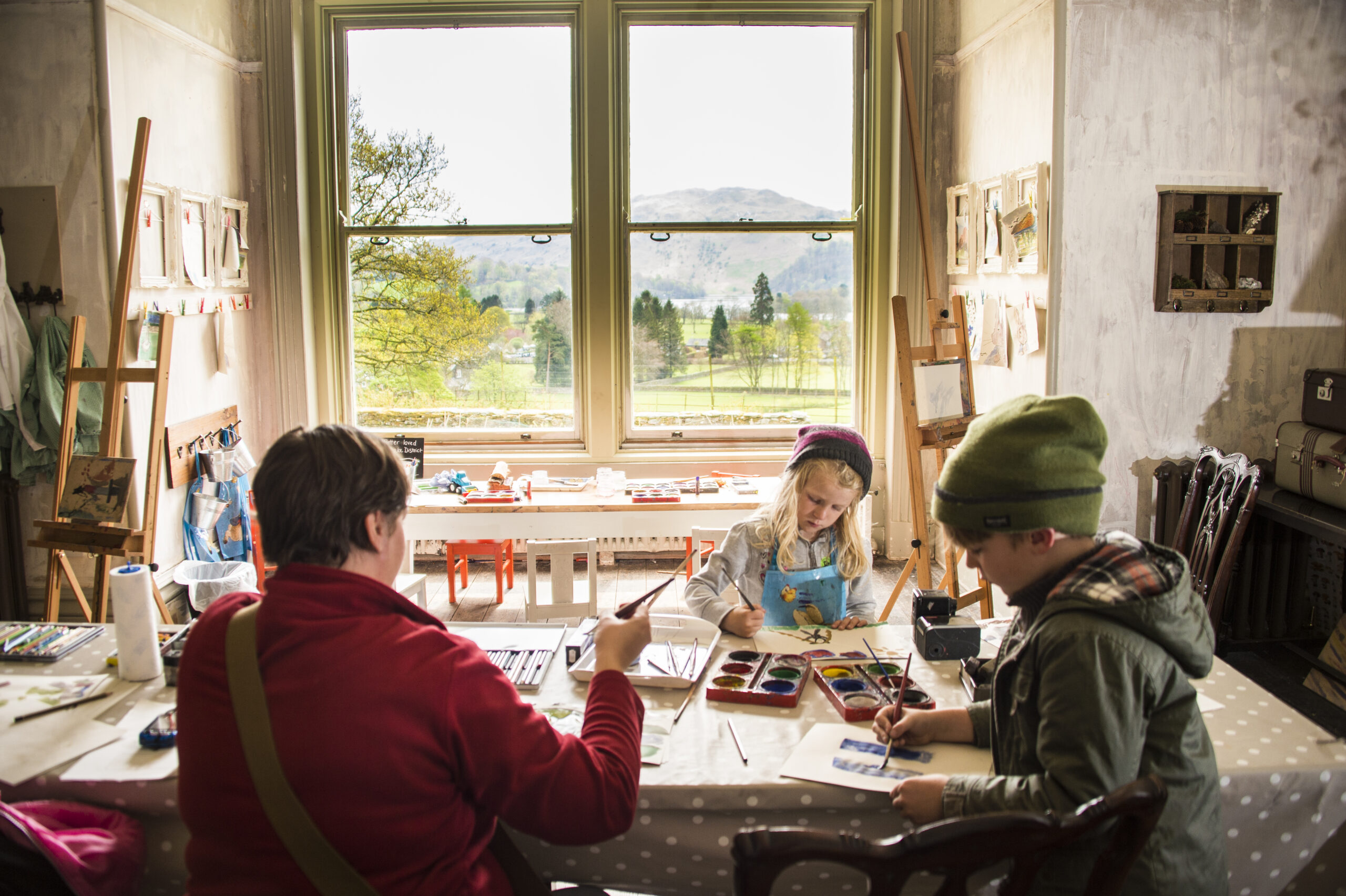 Adult female and two young children sat at a large table painting. Large window with a view over green gardens