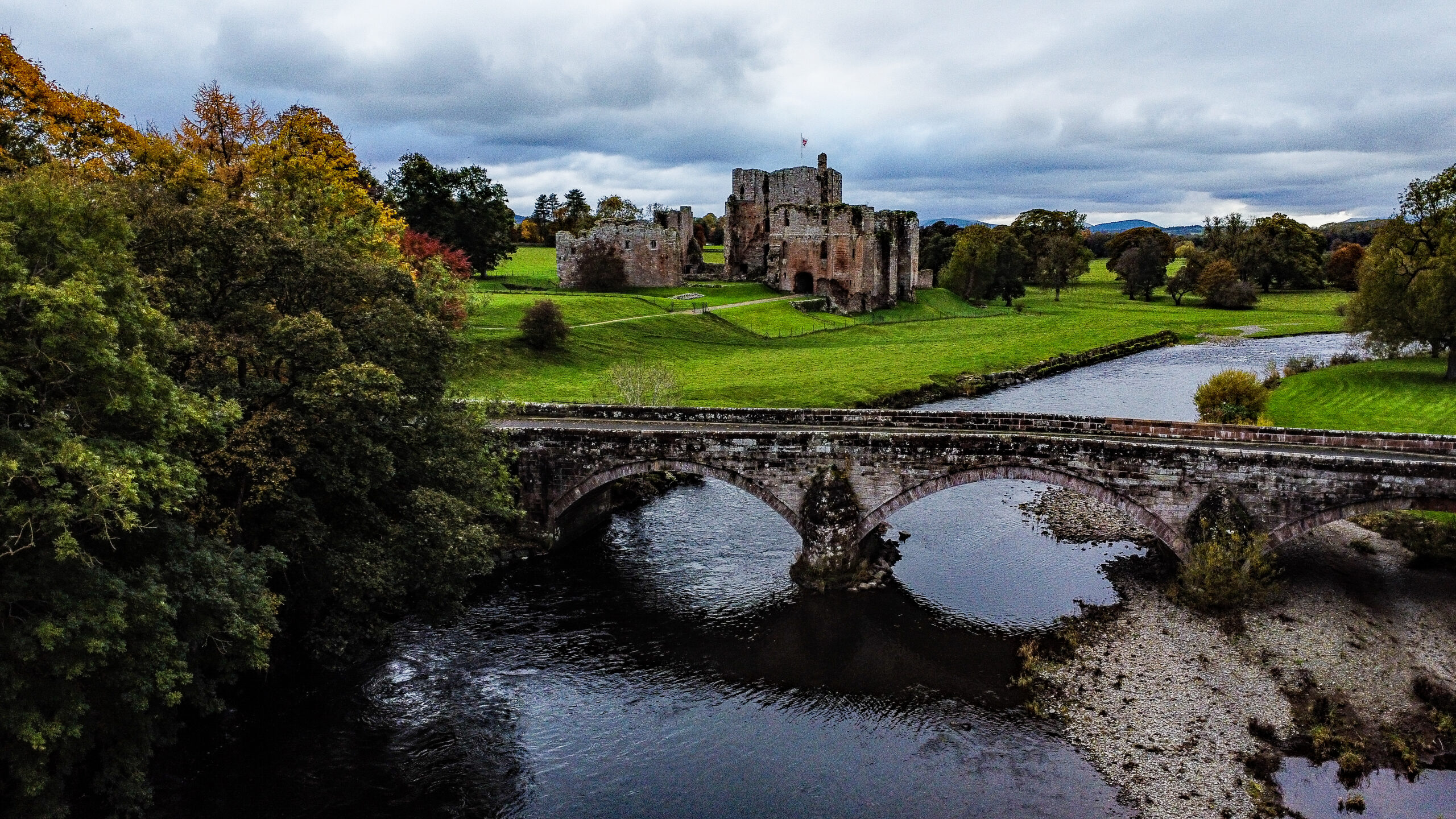Brougham Castle in Penrith. Old stone castle in background, river and stone bridge surrounded by green grass and trees in foreground.