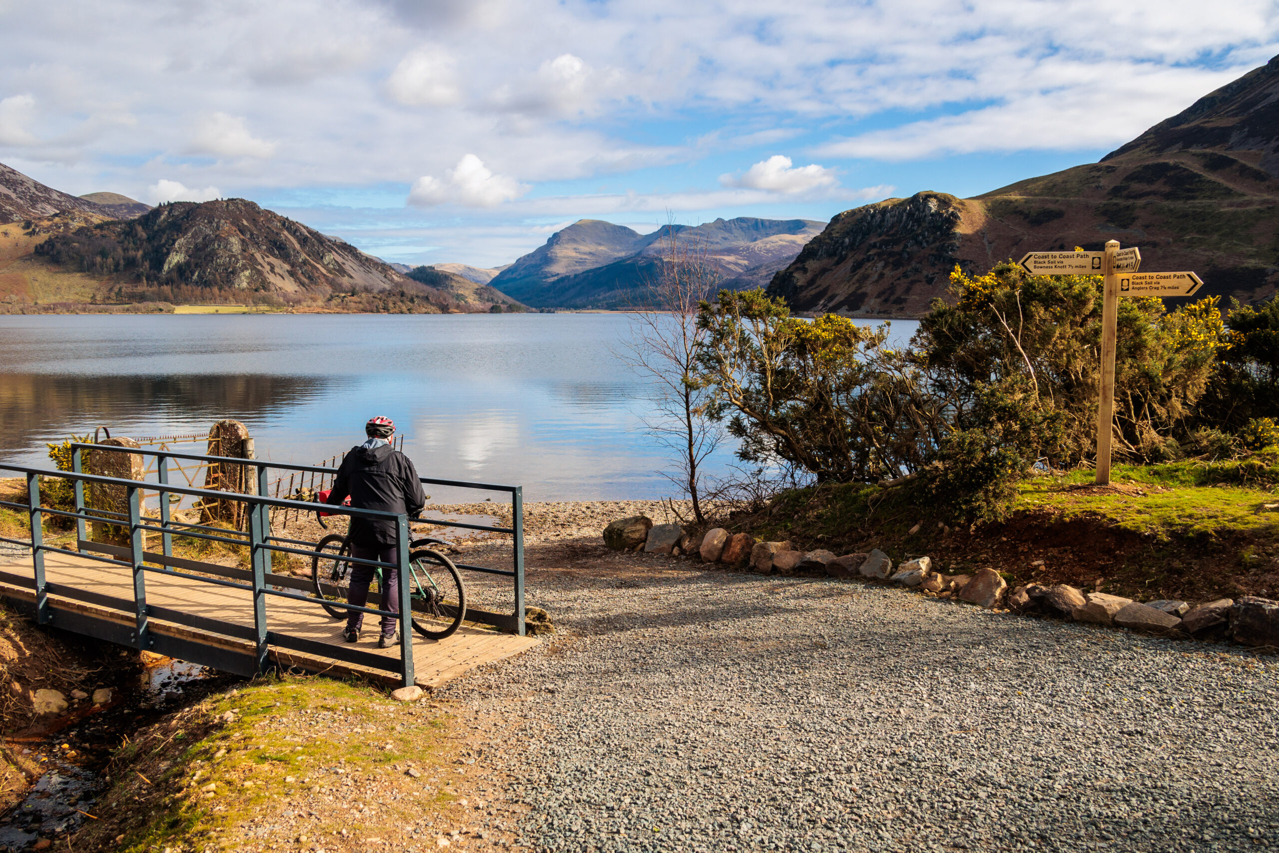 Landscape view of ennerdale water and surrounding fells on a sunny day. Adult male stood with mountain bike on ennerdale bridge