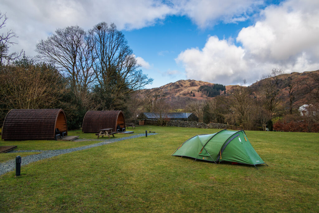 Green field surrounded by brown fells and leafless trees. Small green tent in centre of field next to two wooden camping pods, divided by grey gravel path