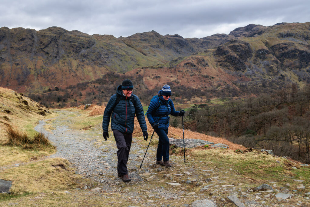 two white adult walkers wearing dark coloured full outdoor kit and walking poles, hiking in autumnal rocky fells