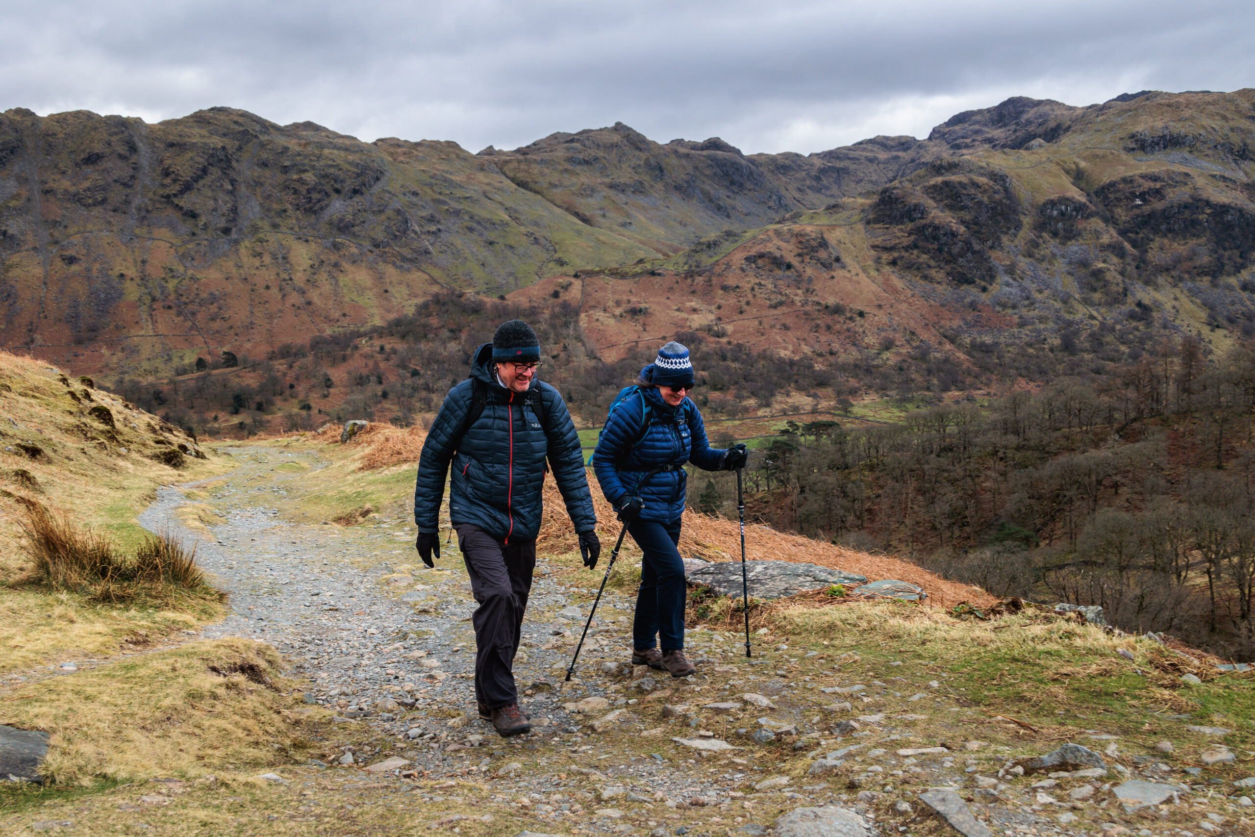 two white adult walkers wearing dark coloured full outdoor kit and walking poles, hiking in autumnal rocky fells