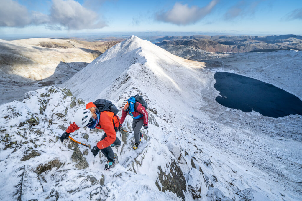 Two people wearing outdoor kit climbing Helvellyn in the snow on a bright day