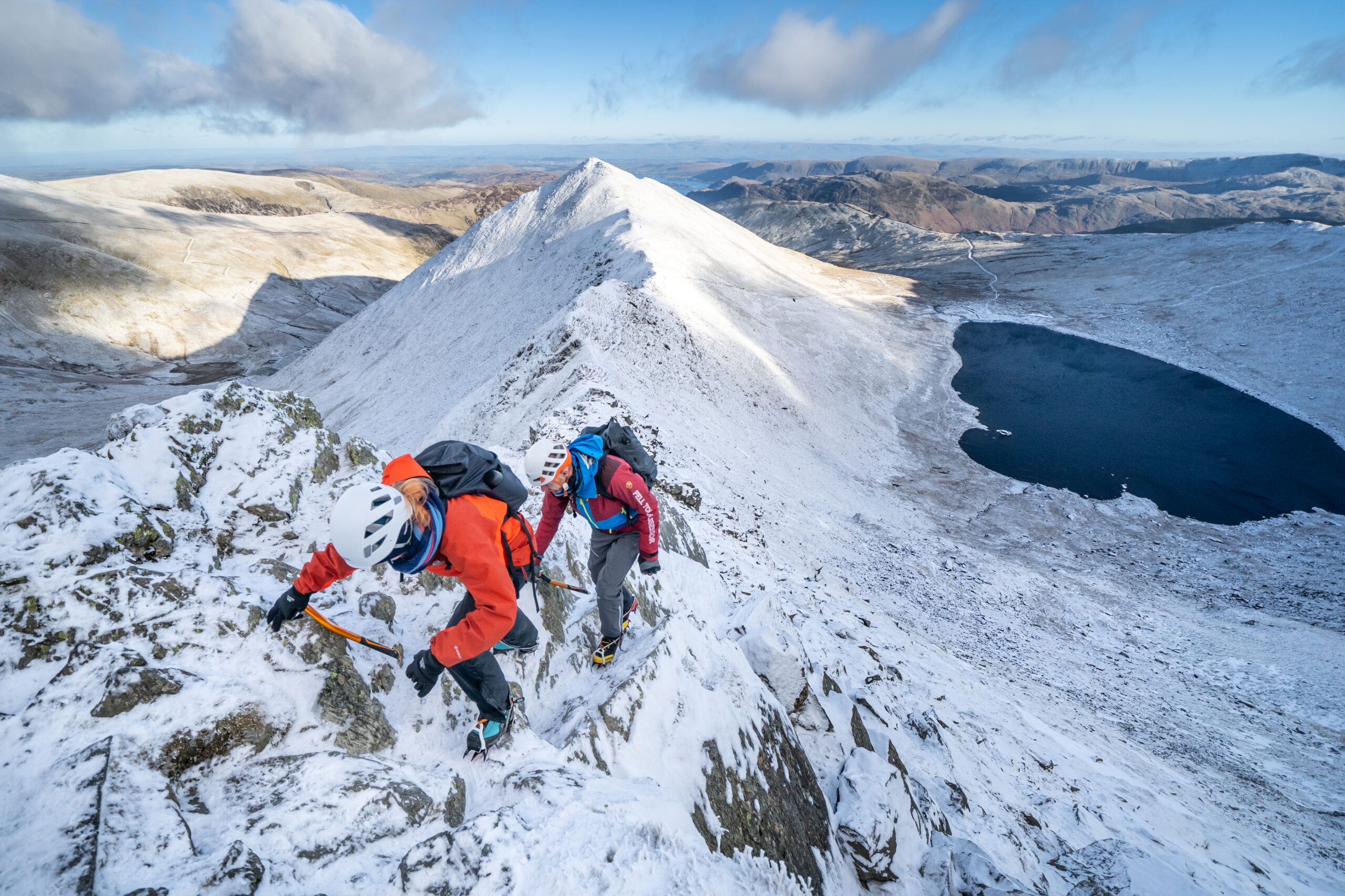 Two people wearing outdoor kit climbing Helvellyn in the snow on a bright day
