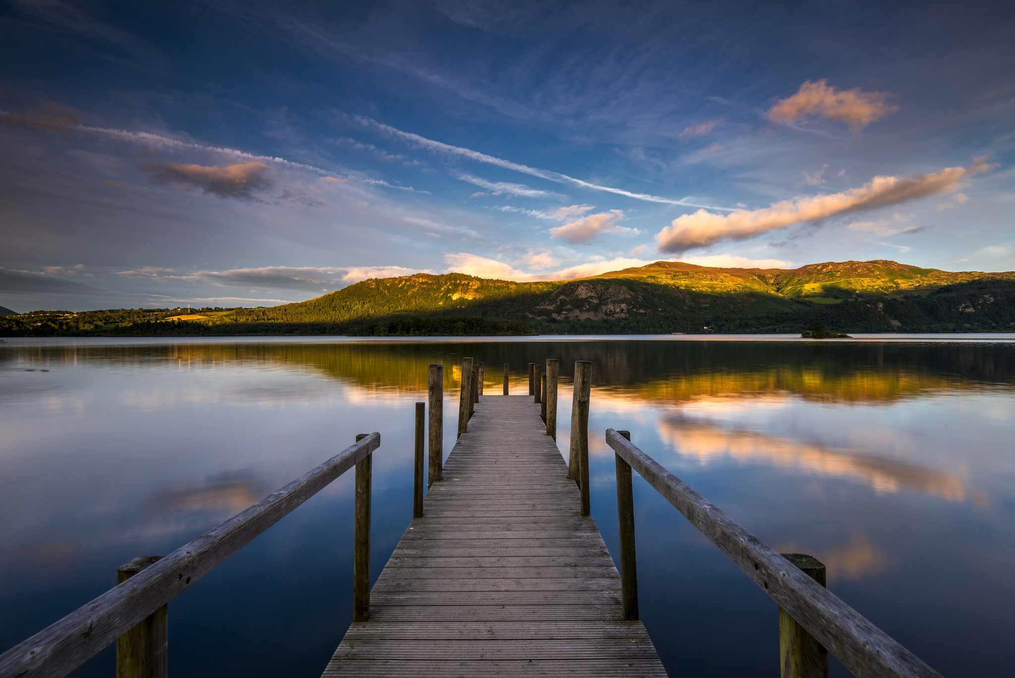 Looking down a wooden jetty into Derwent Water. The lake is blue and orange, reflecting the sunset sky and woodland hills above