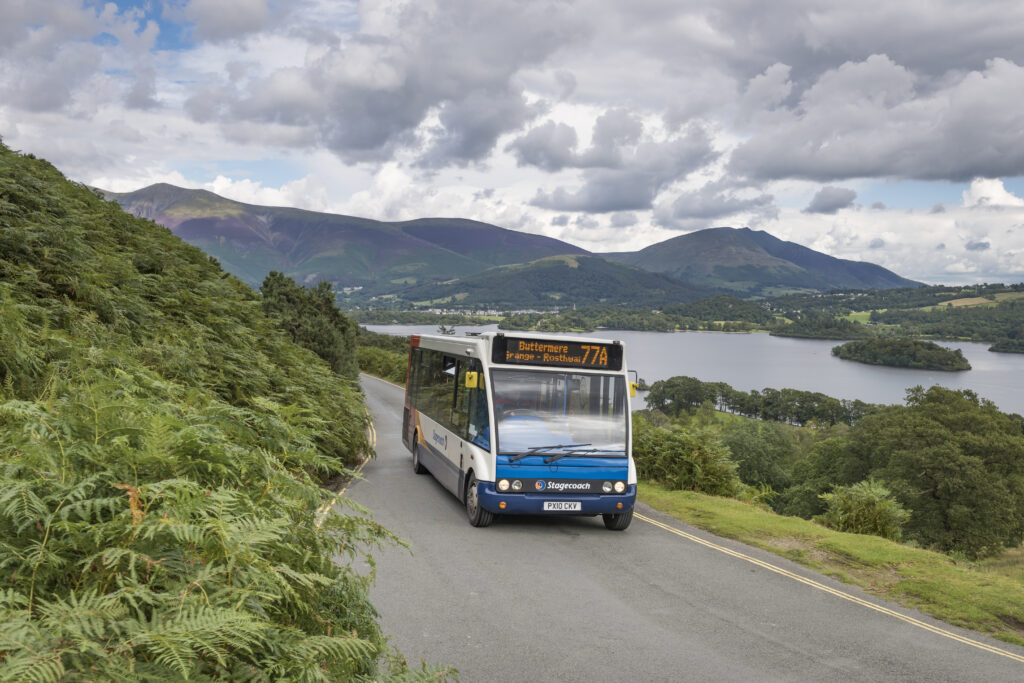 Stagecoach bus travelling along a road alongside lake with fells in the distance