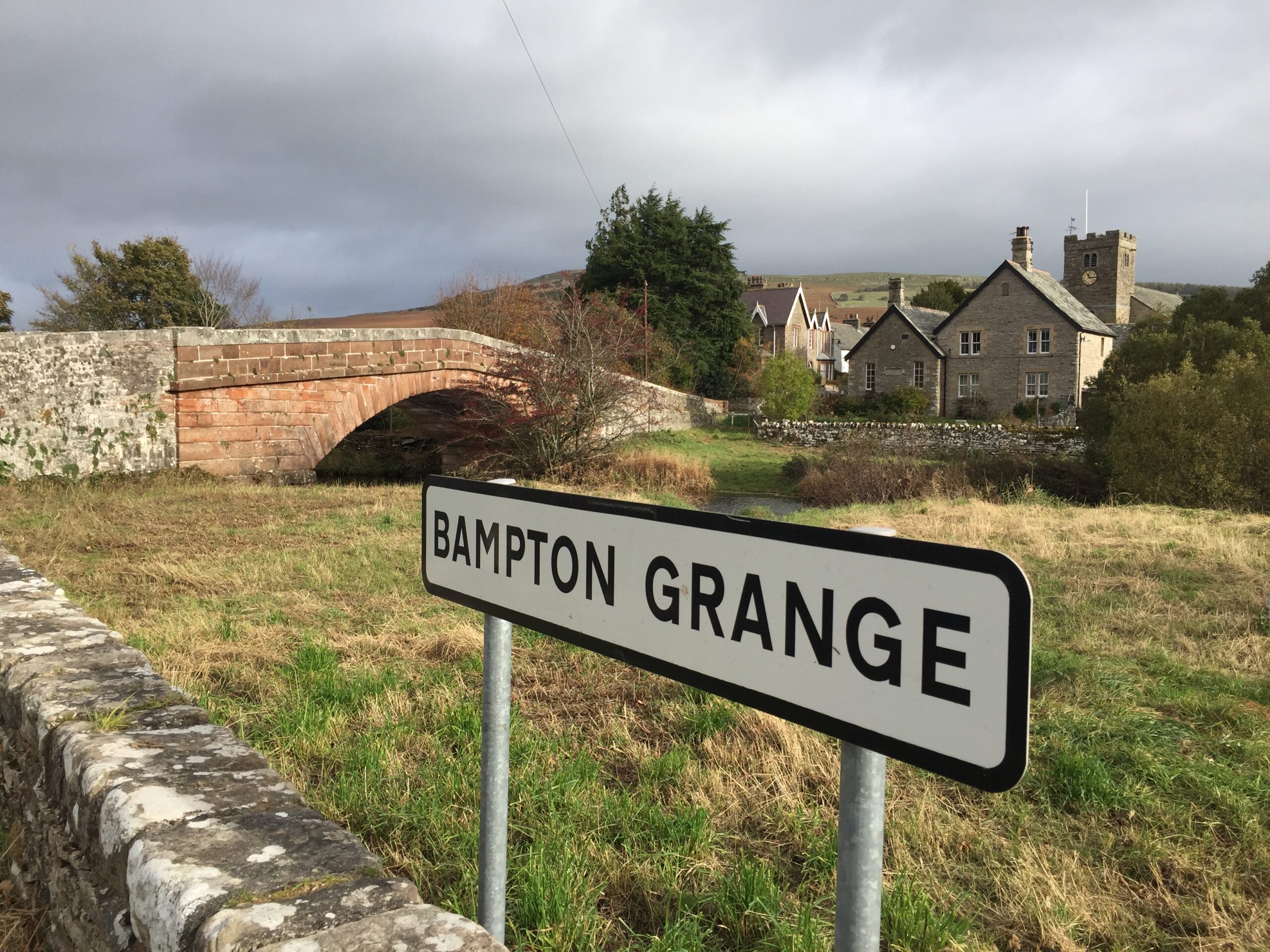 white and black road sign with text reading 'bampton grange.' behind is green and beige grass in front of a red brick bridge and large stone house