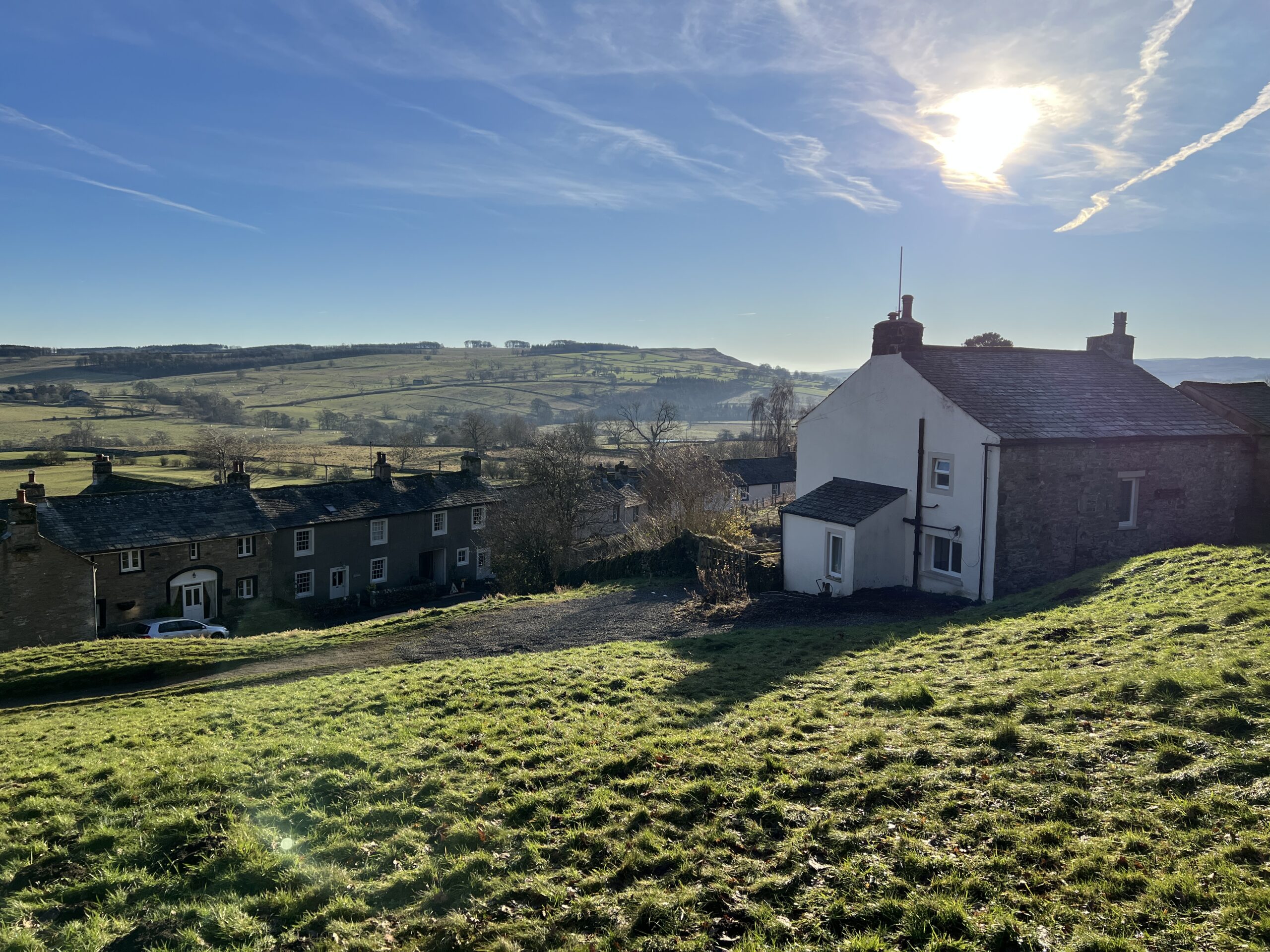 large two story detached white house with slate roof elevated on sloped grass field looking out to rolling green fields and village. blue sky and sunny day
